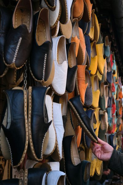 A person holding one shoe in front of a wall full of various colored leather shoes, in the style of Moroccan market raw photography. --ar 85:128