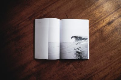 Photo of an open magazine on top, flat lay, on a dark wood table with a white page showing an ocean wave photo, clean and minimal styling, white space around the edge of the picture. --ar 128:85