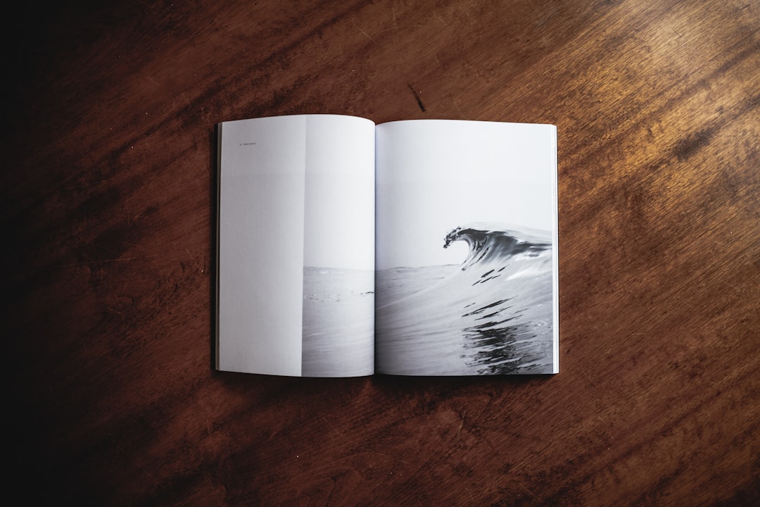 Photo of an open magazine on top, flat lay, on a dark wood table with a white page showing an ocean wave photo, clean and minimal styling, white space around the edge of the picture. –ar 128:85