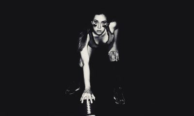 A full body studio photograph of an athletic woman with glasses and long dark hair in her late twenties, kneeling on the ground holding football gloves hands at chest level while wearing black shoes. The background is solid black. She has very thin eyebrows. Her face glows from behind the light source. Black and white photography in the style of Platon photographer, with a grainy film filter. --ar 32:19