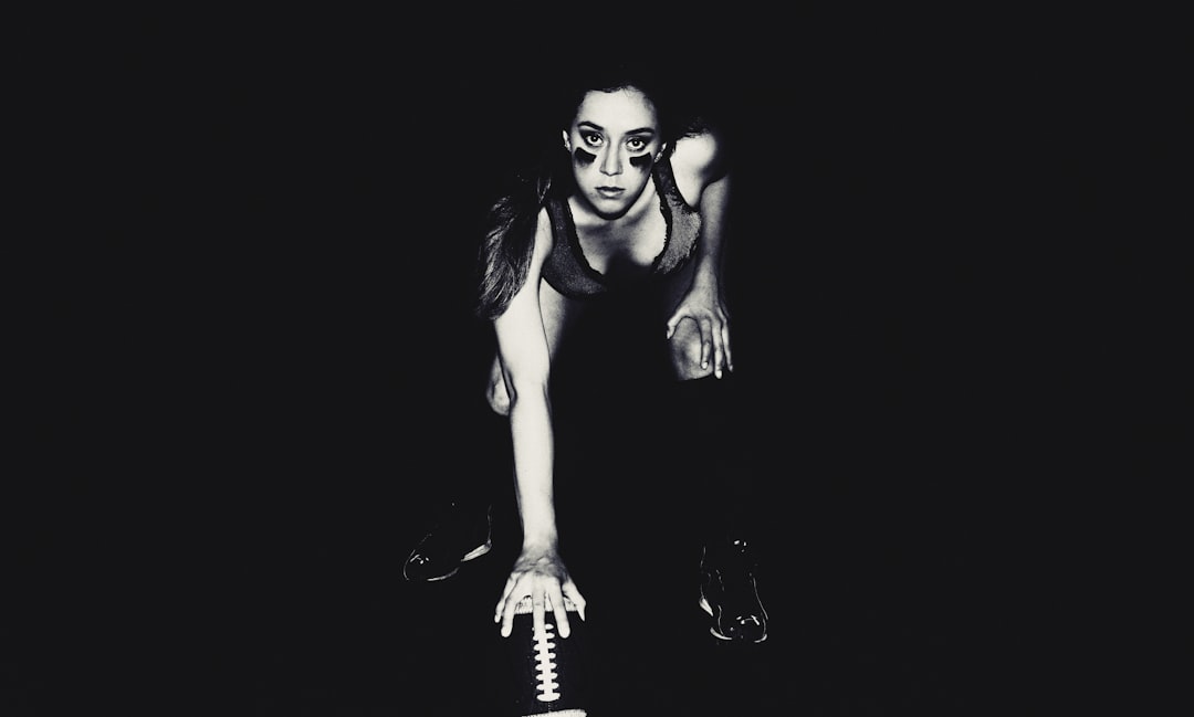 A full body studio photograph of an athletic woman with glasses and long dark hair in her late twenties, kneeling on the ground holding football gloves hands at chest level while wearing black shoes. The background is solid black. She has very thin eyebrows. Her face glows from behind the light source. Black and white photography in the style of Platon photographer, with a grainy film filter. –ar 32:19