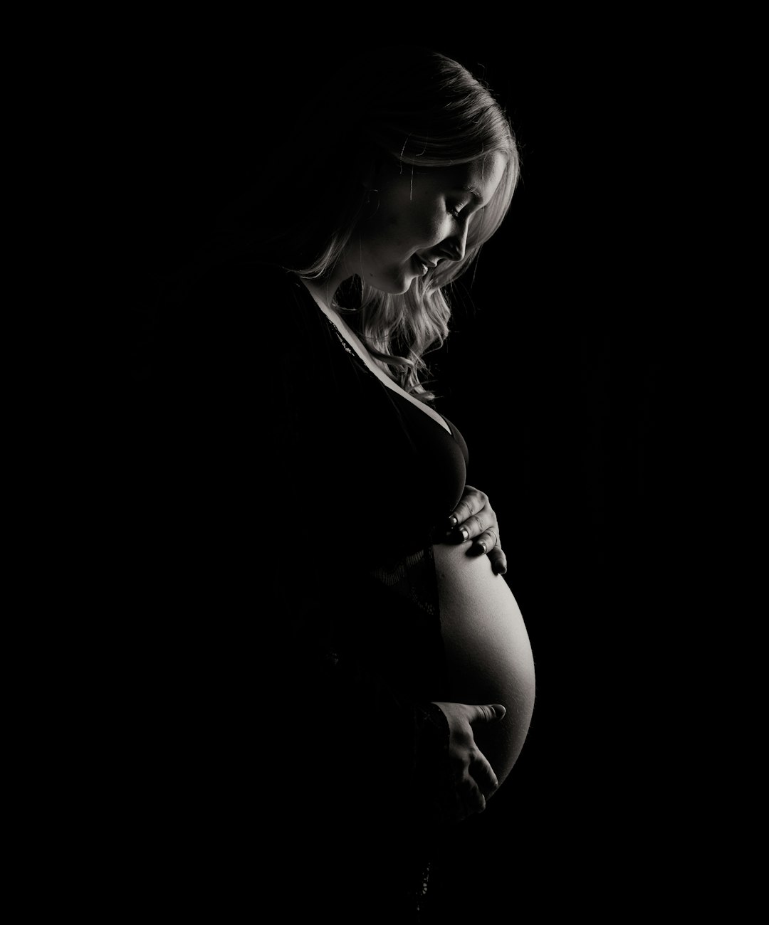 A pregnant woman’s silhouette against the stark black background of her studio, illuminated by soft side lighting that accentuates every curve and detail in the portrait. The focus is on capturing the beauty and elegance of motherhood with the delicate interplay between light and shadow. The photograph has the style of a commercial photo shoot or professional photography. –ar 53:64