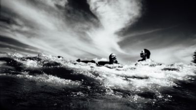 black and white photo of surfers in the ocean, taken from below water level, clouds overhead, low angle, grainy film noir style, wide shot, using a 35mm lens, high contrast, sharp details, dramatic lighting, long exposure, waves splashing up against the camera, in the style of noir films. --ar 16:9