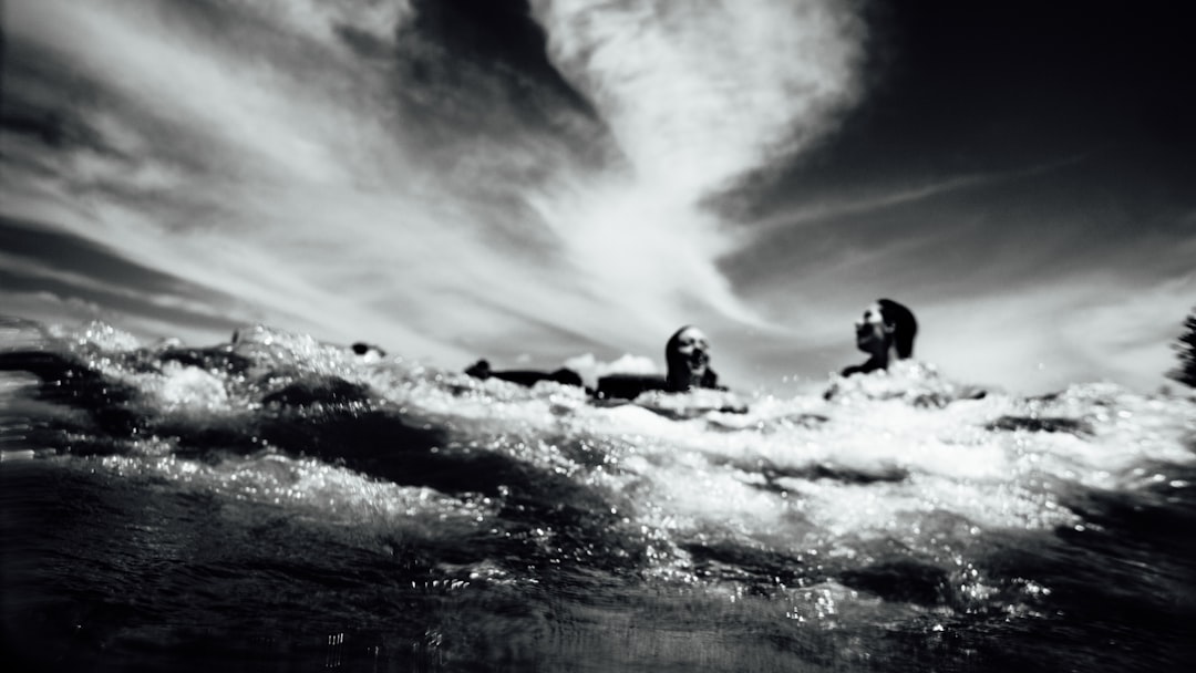 black and white photo of surfers in the ocean, taken from below water level, clouds overhead, low angle, grainy film noir style, wide shot, using a 35mm lens, high contrast, sharp details, dramatic lighting, long exposure, waves splashing up against the camera, in the style of noir films. –ar 16:9