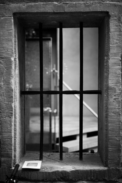 Black and white photography of an iron window with bars, centered in the frame. The view outside is a stairway to another building. --ar 85:128