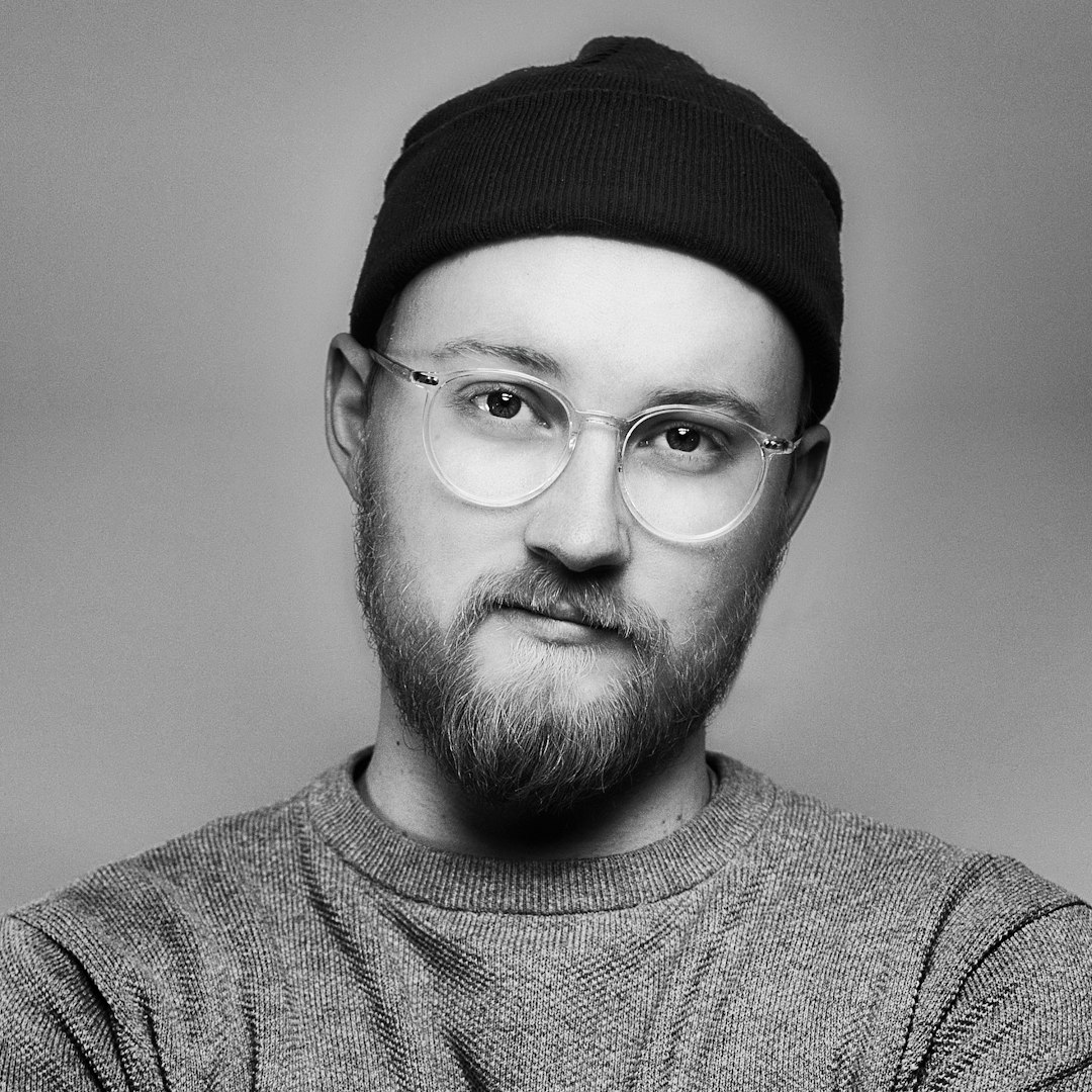 Portrait of a white man with a beard, wearing glasses and a black beanie, looking at the camera, grey sweater, in the style of a studio portrait, high contrast between foreground and background, minimalistic, award-winning photography, simple studio background, quirky, shot with a Hasselblad H6D400C MultiShot camera from multiple angles.