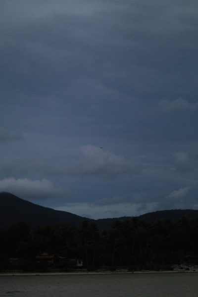 Dark grey clouds covered the sky over Saho harbor, and mountains could be seen in the distance. The photo was taken from near sea level with trees visible on both sides of the beach. A distant airplane was flying overhead. In front of the camera there was one house and a small boat docked at the shoreline. The image had a dark blue tone. It showed a calm ocean with gentle waves and subtle ripples on the water surface. --ar 85:128