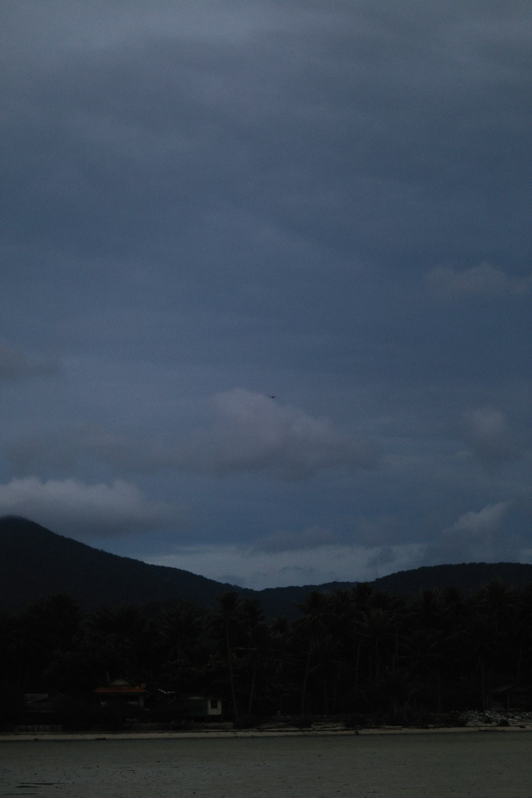 Dark grey clouds covered the sky over Saho harbor, and mountains could be seen in the distance. The photo was taken from near sea level with trees visible on both sides of the beach. A distant airplane was flying overhead. In front of the camera there was one house and a small boat docked at the shoreline. The image had a dark blue tone. It showed a calm ocean with gentle waves and subtle ripples on the water surface. –ar 85:128