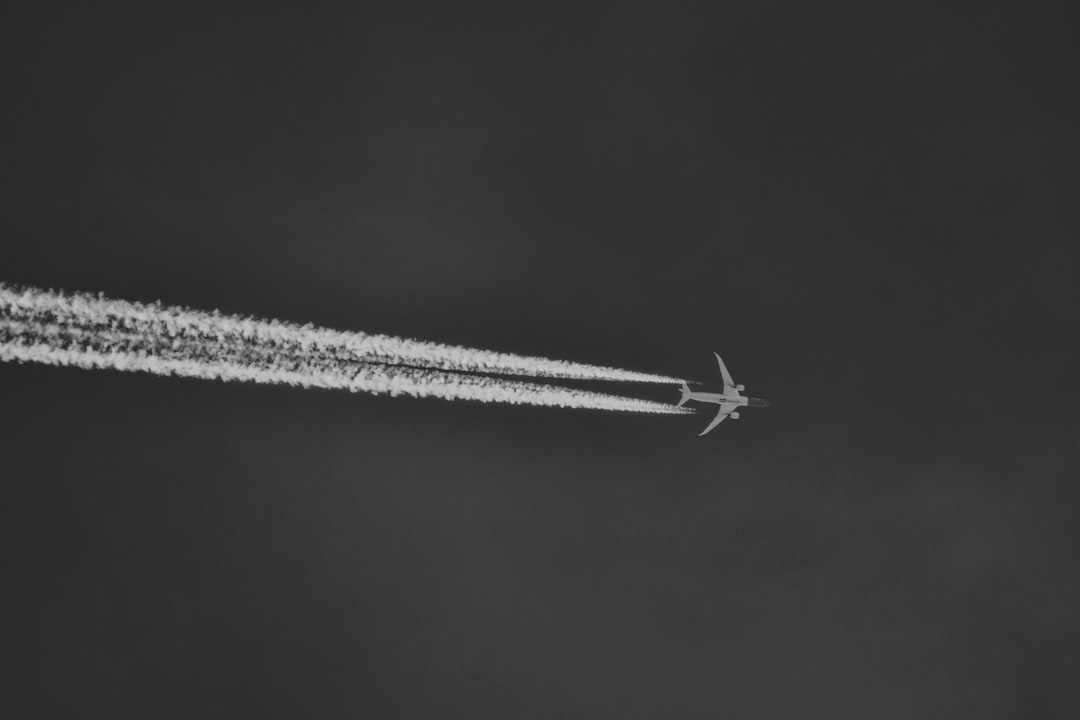 A grayscale photo of an airplane flying in the sky with its trail visible, symbolizing travel and journeying. The plane is seen from below against a clear blue or grey background. The photo is in the style of an anonymous artist. –ar 128:85