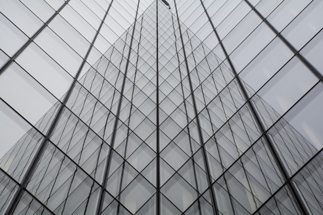 A glass building with diagonal patterned windows, creating an abstract and modern appearance. The building’s tall walls feature symmetrical geometric shapes that create depth in the photograph. A person is seen climbing up one of these vertical panels, adding a human element to the architecture. –ar 128:85