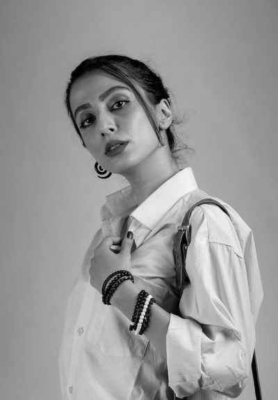 A black and white studio portrait of an Indian woman in her mid20s, wearing oversized men's shirt with sleeves rolled up to the shoulder line, holding handbag over one arm, posing for fashion editorial photoshoot. She has hair tied back in low ponytail hairstyle, hoop earrings, pearl bracelets on each wrist. Her pose is confident but playful, looking at camera, with soft lighting that highlights details like texture or pattern of . The background should be simple and clean, focusing attention on subject --ar 11:16
