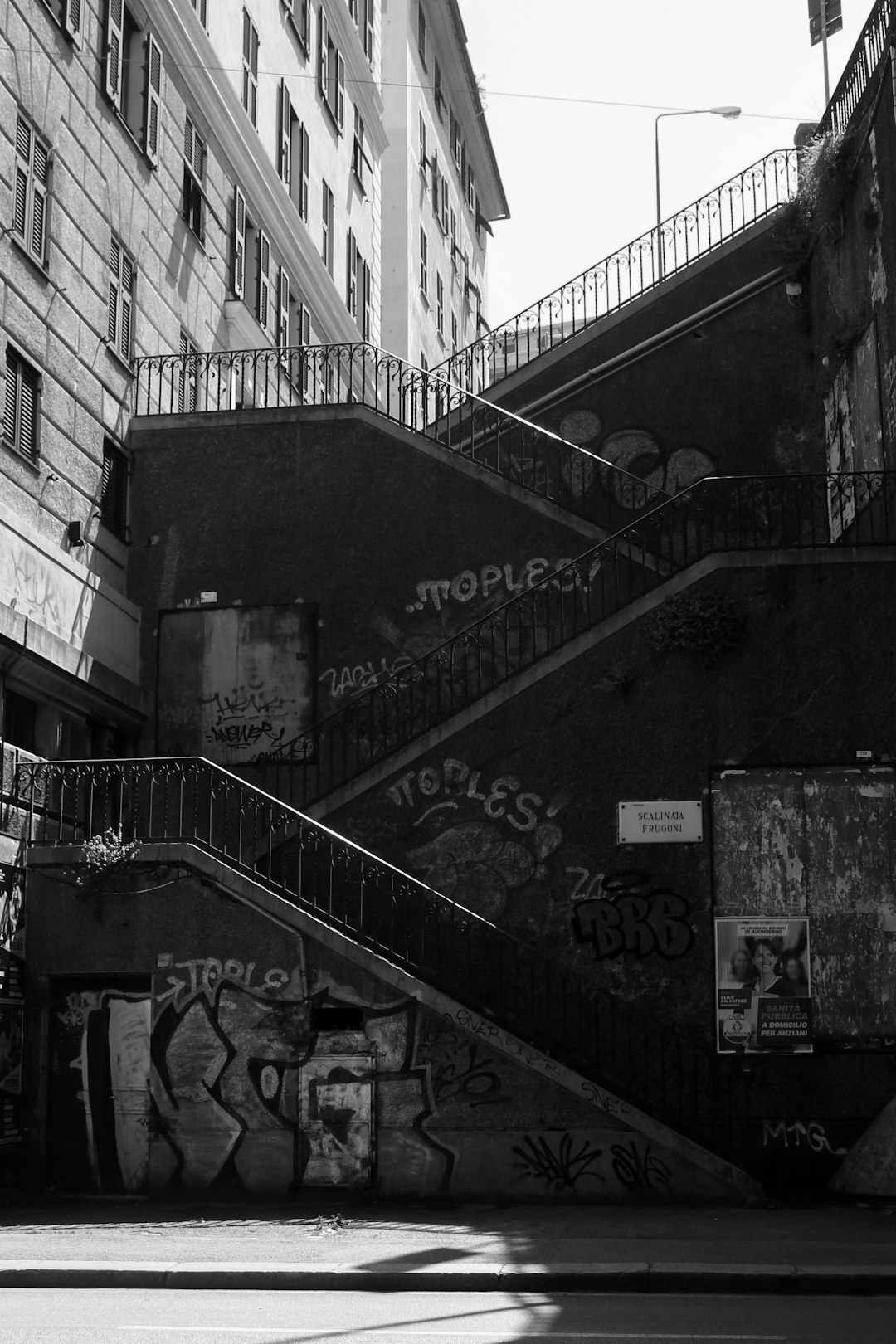 A black and white photo of the stairs leading up to an urban street in Naples, Italy with graffiti on them. In front there is another set of steps leading down into the streets below. The city has a dark aesthetic. There are some shadows from trees and buildings. Shot in the style of 35mm film grain Kodak photography, in the style of Fujifilm superia Venus 800. –ar 85:128