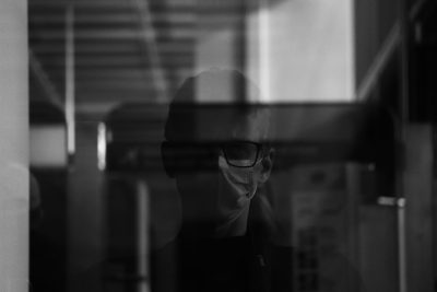 A scientist wearing a protective mask is seen through the window of an office, blurred in black and white. The camera captures him from behind as he walks past his desk with glass panes. He has short hair tied back to reveal glasses on top of it. His face peeks out between two windows that frame the scene, creating a dark atmosphere. A shadowy figure stands in the style of another door at the right side of the picture. The shot was taken from inside looking outside. --ar 128:85