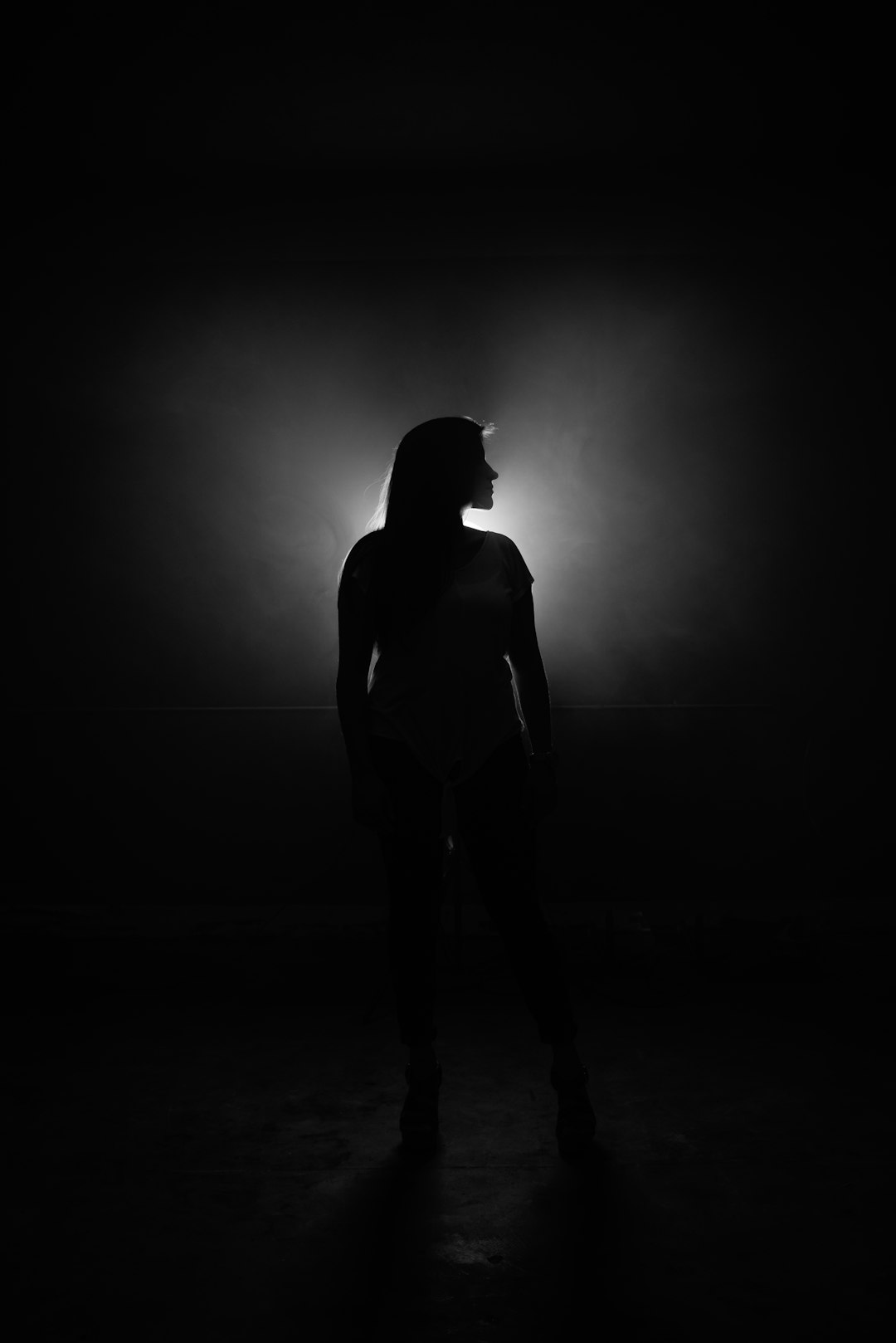 A black and white photograph of an athletic woman’s silhouette against the darkness, illuminated by soft light from above. The background is dark with subtle lighting effects creating a dramatic effect. She stands in front of a simple backdrop, wearing high heels that add to her height., focus on face –ar 85:128