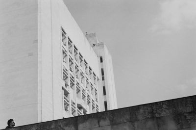 Black and white photography of an art deco building in Rio de Janeiro, Brazil. A man is hanging from the wall looking up at it in the style of. --ar 32:21