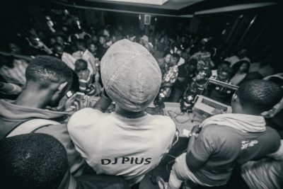 A crowd of people wearing white t-shirts with "DJplus" written on the back, standing in front of a DJ playing music at an African night club party. Black and white photography. The crowd watches the DJ playing music in the style of DJplus. --ar 128:85