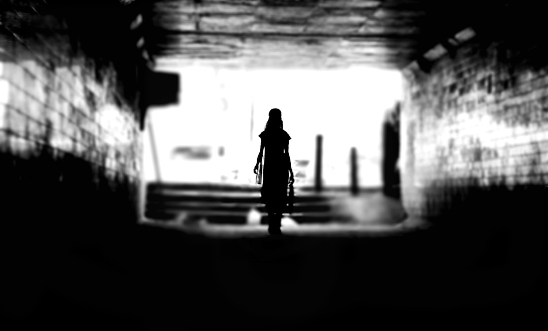 Silhouette of a woman standing in a dark tunnel, with low light and a distant view against a white background, with black and grey tones and high contrast, captured with a wideangle lens and soft side lighting, creating a contemplative mood. In the distance is an empty street with only one person walking towards her, wearing a long dress and holding a sword. The scene has an eerie atmosphere, as if she were about to face something dangerous or mysterious. Her posture reflects fear but also determination. Done in the style of black and white photography. –ar 128:77