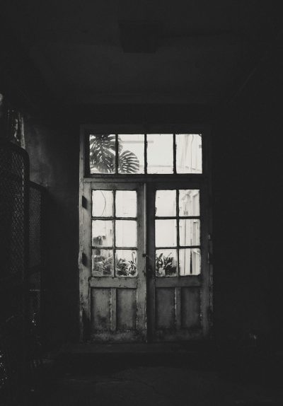 a black and white photograph of an old wooden door with large glass windows, inside the house there is a small window in front of it through which you can see plants outside, the photo was taken from behind so that we only have half a wall visible, the rest has dark shadows, the atmosphere looks gloomy and sad, the background is blurry and dark --ar 89:128