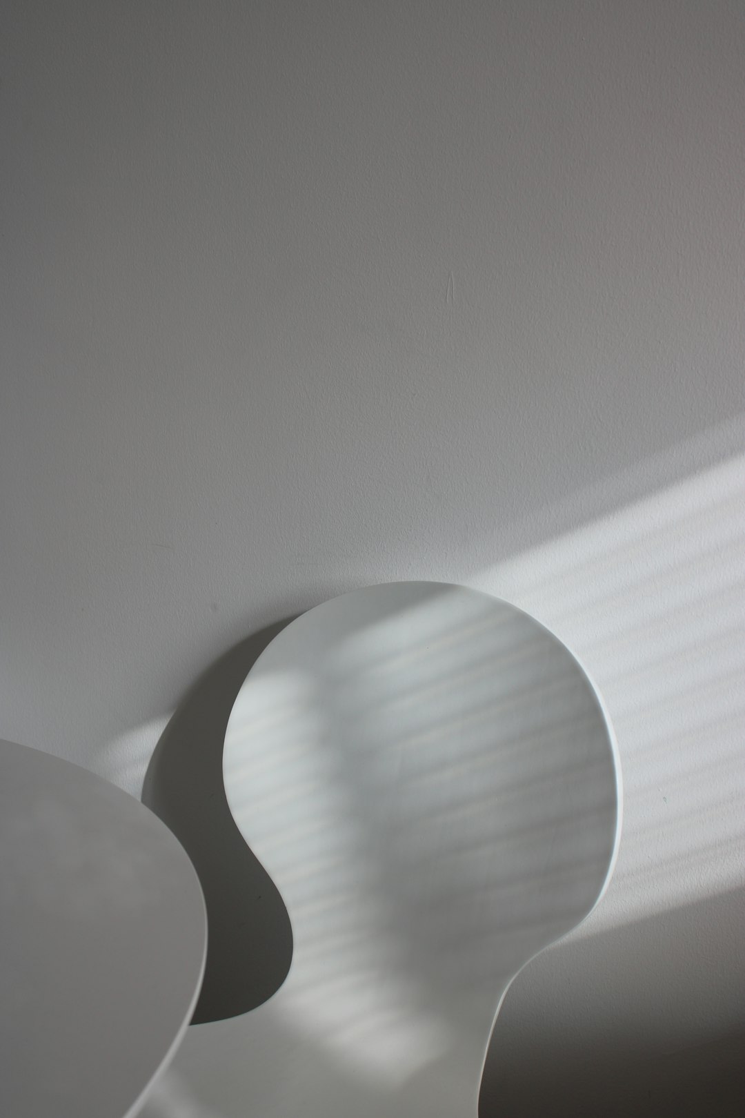 A closeup of the white wall, with two circular shadows cast on it by an abstract shape. The shadow is a semicircular shape that forms an organic curve, with its edges slightly blurred and textured. It creates a sense of lightness through the interplay between sunlight and shadows. This minimalist composition captures the essence of nature’s tranquility in a contemporary setting. –ar 85:128