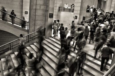 A black and white photograph of the British Museum, busy with visitors moving up the stairs in blurry motion, was captured from above. The focus is on one individual standing out among others due to their distinct appearance or posture. --ar 128:85