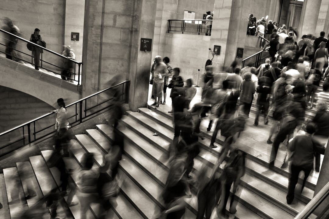 A black and white photograph of the British Museum, busy with visitors moving up the stairs in blurry motion, was captured from above. The focus is on one individual standing out among others due to their distinct appearance or posture. –ar 128:85