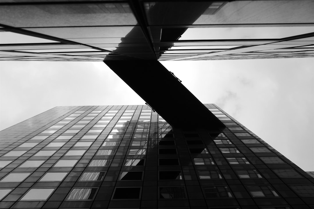 A black and white photo shows the outside corner wall of an office building, with reflections on glass windows. The perspective is looking up from below at sky level. There is no human presence in the frame. In the front, another tall skyscraper is visible through the window, in the style of a photo taken with a Sony Alpha A7 III camera and a wide-angle lens. –ar 128:85