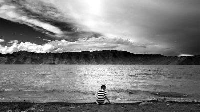 Black and white photography of an Asian man wearing striped prison  sitting on the shore at Lake dinosaur, looking out over the lake. The clouds overhead paint beautiful skies. He is alone with his thoughts as he sits by the water’s edge. His gaze was fixed upon its surface, reflecting everything around it in the style of China's Loess Hathight, with mountains behind him. --ar 16:9