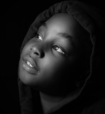 Black and white studio portrait of an African American teen girl wearing a hoodie, looking up with light in her eyes, high contrast, high resolution, high detail, taken with an 80mm lens at f/2.4, cinematic style with global illumination, in the style of no specific artist. --ar 59:64