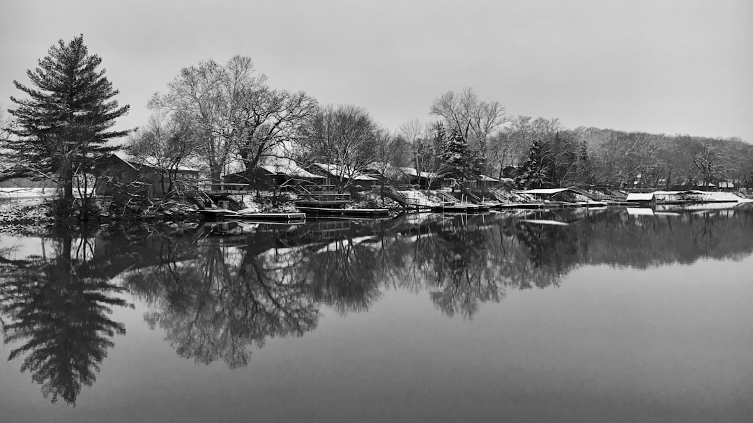 Black and white photo of the reflection in the water, small village houses along the river bank, trees, winter landscape, snow covered ground, trees with no leaves, trees reflecting on the calm body of water, snowy forested hills behind the houses, river flowing through town, snowy sky. –ar 16:9