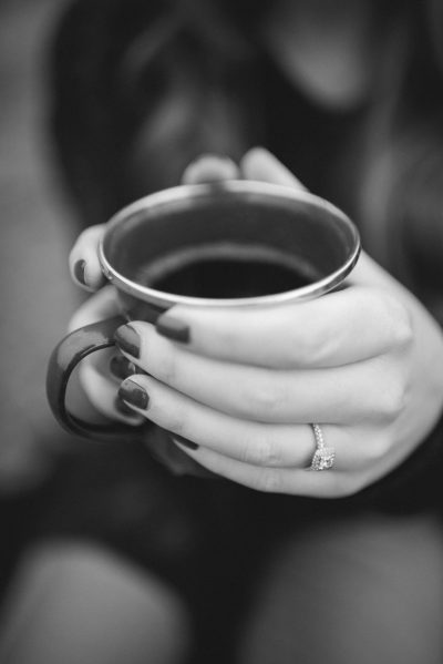 Black and white photography of a female's hands holding a coffee mug with an engagement ring on it, in a minimalistic setting. --ar 85:128