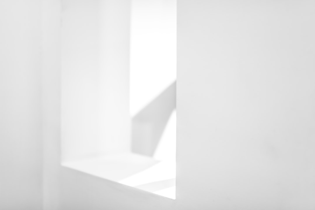 White background with a small window or door casting soft shadows, creating an abstract and minimalist composition. The light from the open doorway creates gentle highlights on the white surface below, adding depth to the scene. –ar 128:85