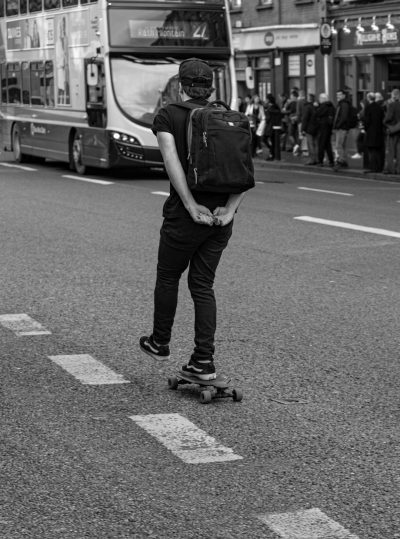 A black and white photo of someone skateboarding down the street in Dublin. An Irish young man with a cap on his head, wearing a backpack, riding a skateboard on the road next to a double decker bus. A crowd watching him. Street photography with a depth of field capturing street style and street life scenes in the style of street photography. --ar 95:128