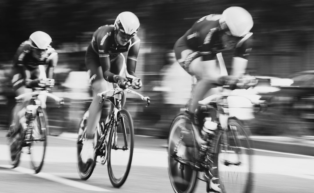 Black and white photography of three professional cyclists during a time trial race in competition, wearing black jerseys with their team name on the back. They have helmets for protection while riding their bikes at high speed through city streets. The scene captures them speeding past blurred spectators, creating an atmosphere of intense racing action. In sharp focus. –ar 64:39