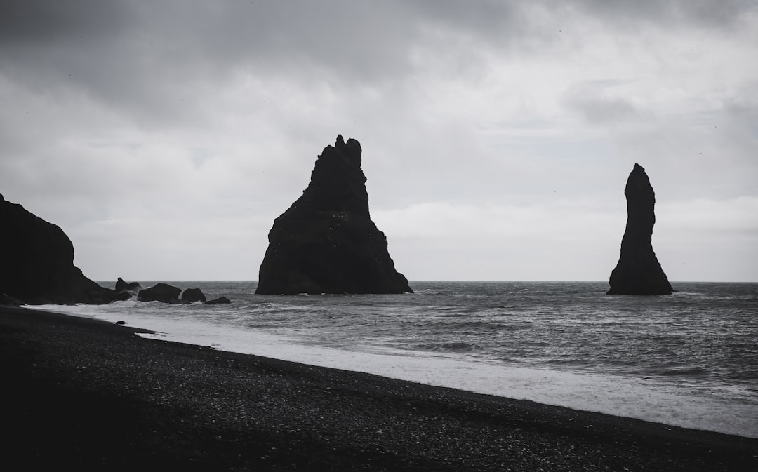 Black rock spires rising from the sea, dark sand beach, Iceland, overcast sky, wide angle, black and white photography, film grain, cinematic, hd, no contrast, no sharpness, soft focus, moody light, mood lighting, –ar 128:79