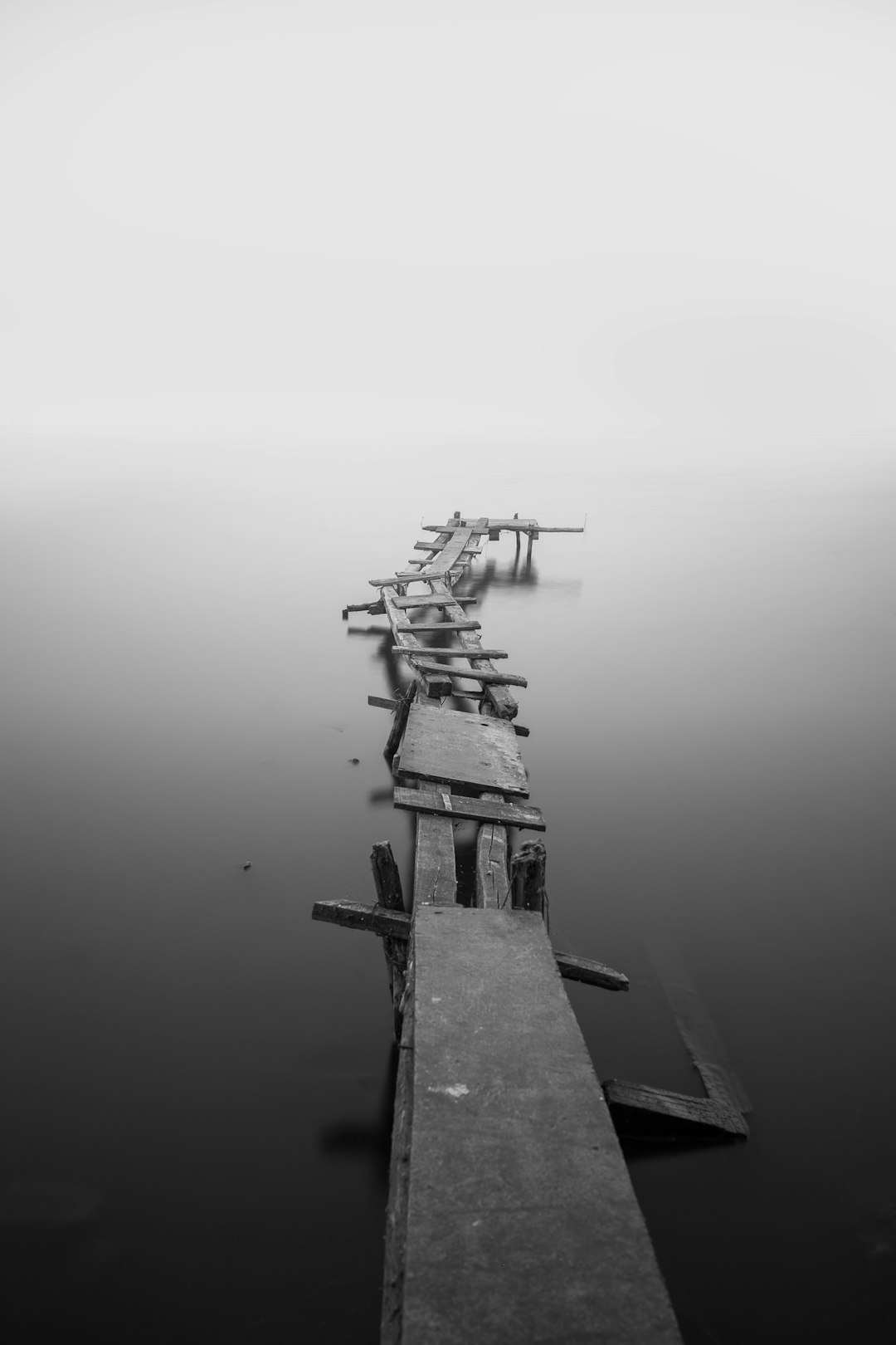 black and white photo of a broken wooden bridge over calm water on a foggy morning, long exposure, shot on a Sony Alpha A7 III with an f/8 lens in the style of no artist. –ar 85:128