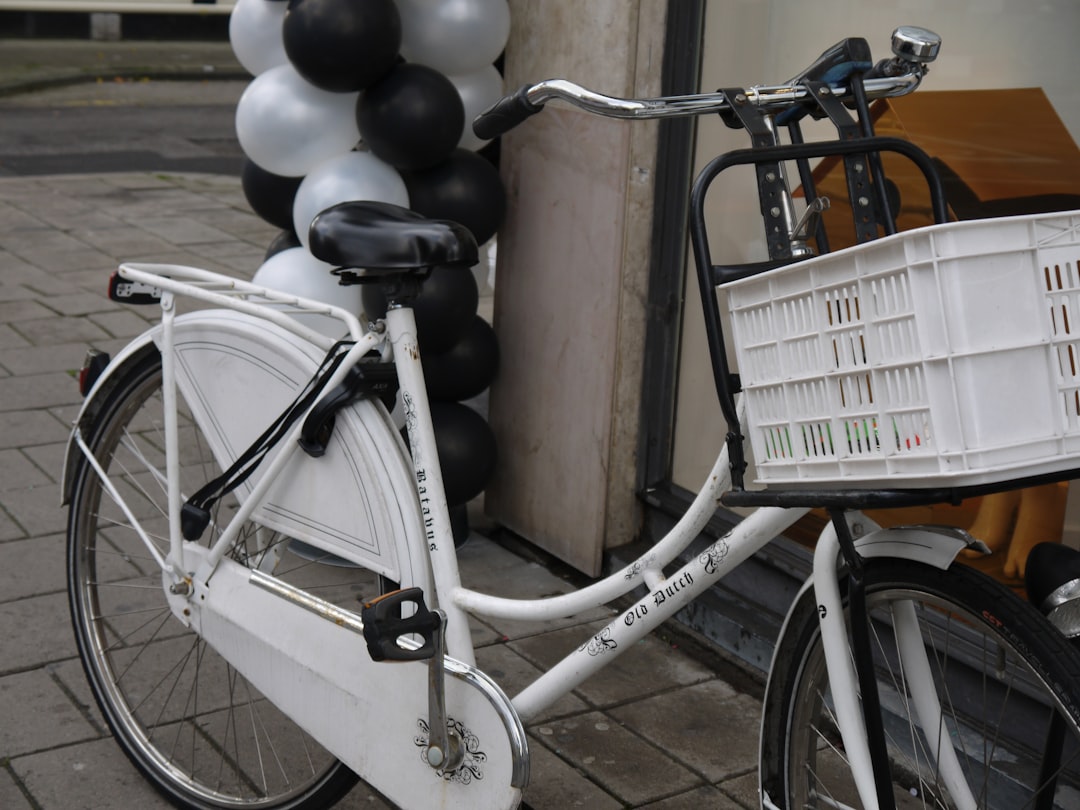 A white bike with black and silver accents, carrying an empty plastic crate on the front basket. It’s parked outside of Amano chocolate shop in Amsterdam which has grey walls and is decorated with lots of black balloons. The store name “Banium” can be seen painted onto one wall. –ar 4:3