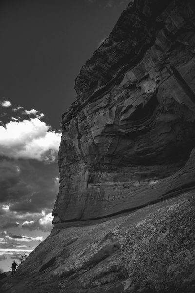 Black and white photograph of the side profile of Moab's sandstone rock face. A person is riding up to it on horseback in a wide angle shot landscape photography taken with a Nikon Z7 II and an f/2 lens, creating dramatic contrast between highlights and shadows, emphasizing texture in the rocks and sky and conveying a sense of adventure and exploration. --ar 85:128