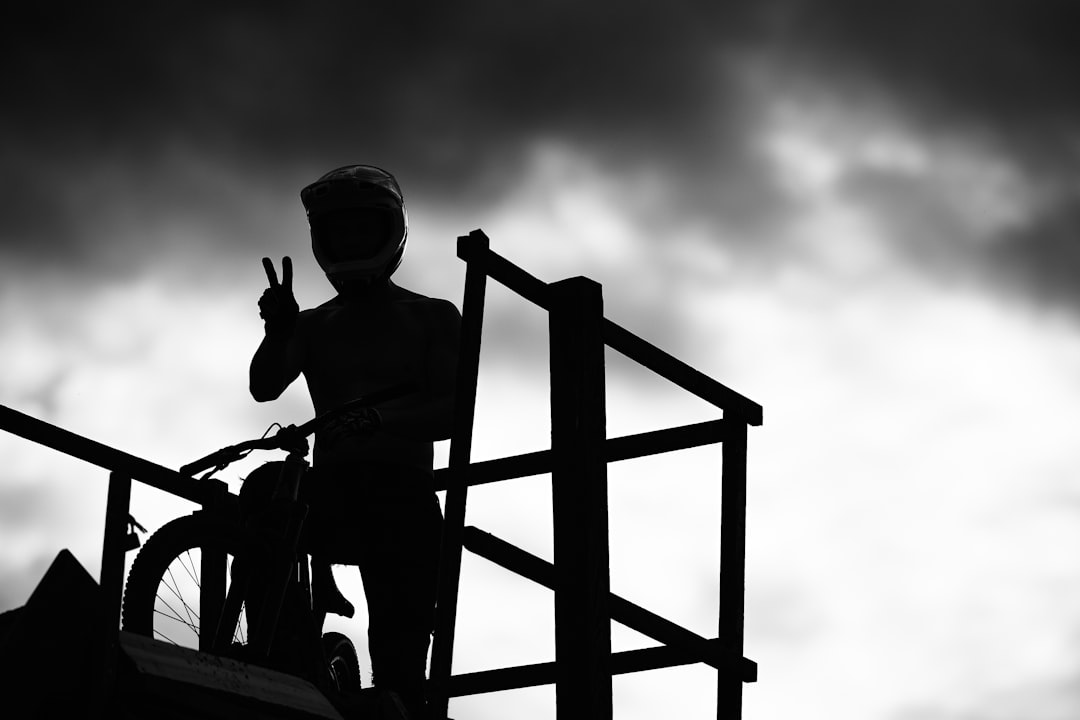 black and white silhouette of an un baclqued biker with helmet standing on top the roof in smFed evil mood showing peace sign, at parkour playground with metal stairs and wooden fence, sky is dark clouds, cinematic shot, Sony Alpha A7 III –ar 128:85