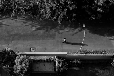 A man running on the road, black and white photography, top view, high angle shot, from above, a park in the background, trees, daylight, sunlight, shadows, a concrete bridge, the long shadow of an old woman sitting under a street light pole, in the style of canon eos r5. --ar 128:85