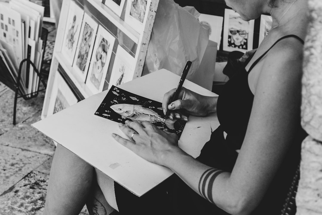 Black and white street photography shows a woman drawing on a blank canvas as she sits outside an art market. In front of her are multiple art prints for sale. Her hand holds a pen with ink that she uses to draw on the canvas. The focus point should be her hands and what is being drawn on the paper, while the background is blurred out. –ar 128:85