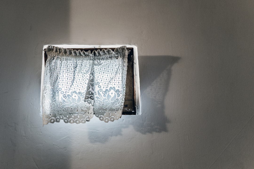 A small square window with lace curtains on the wall, casting long shadows on the white walls. The photography style is simple in composition with a delicate texture of lace curtain and soft lighting that shows delicate details and warmth. –ar 128:85