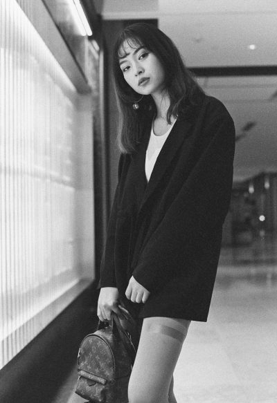 A young Chinese woman wearing an oversized blazer, white tights and a Louis Vuitton monogram baggy short in the airport lobby, photographed in the style of Wong Kar Wai in black & white film photography with a grainy effect. --ar 87:128