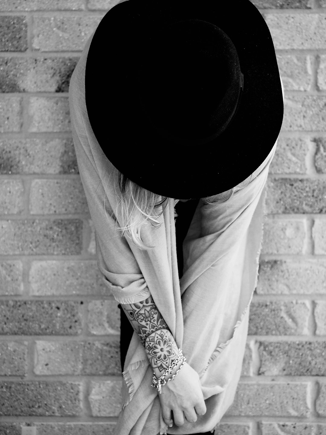 Black and white street photography of an elegant woman with tattoos covering her arm, wearing a black hat on top of the scarf that she is wrapped around to cover up most of it’s face, standing in front of a brick wall, looking down at one hand which holds onto his pants pocket. –ar 3:4