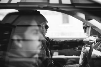 Black and white photo of a man in a beanie sitting inside a car, holding the steering wheel with his hand, with a reflection on the windshield, shot from behind the driver's seat, in an urban setting, with a gritty style in the style of Leica Q2 Monochrom, with an f/4 lens for sharp focus on the subject and a soft background bokeh effect. --ar 128:85