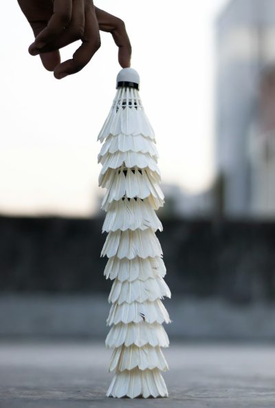 a hand holding badminton shuttlecock, the head of which is made up by many white feathers, stacked on top each other in vertical line to form a mini tower that stands at an angle on its base. The background shows part of a building and sky. The focus is on capturing details such as texture of feather. The overall mood evokes tranquility with minimalism. --ar 43:64