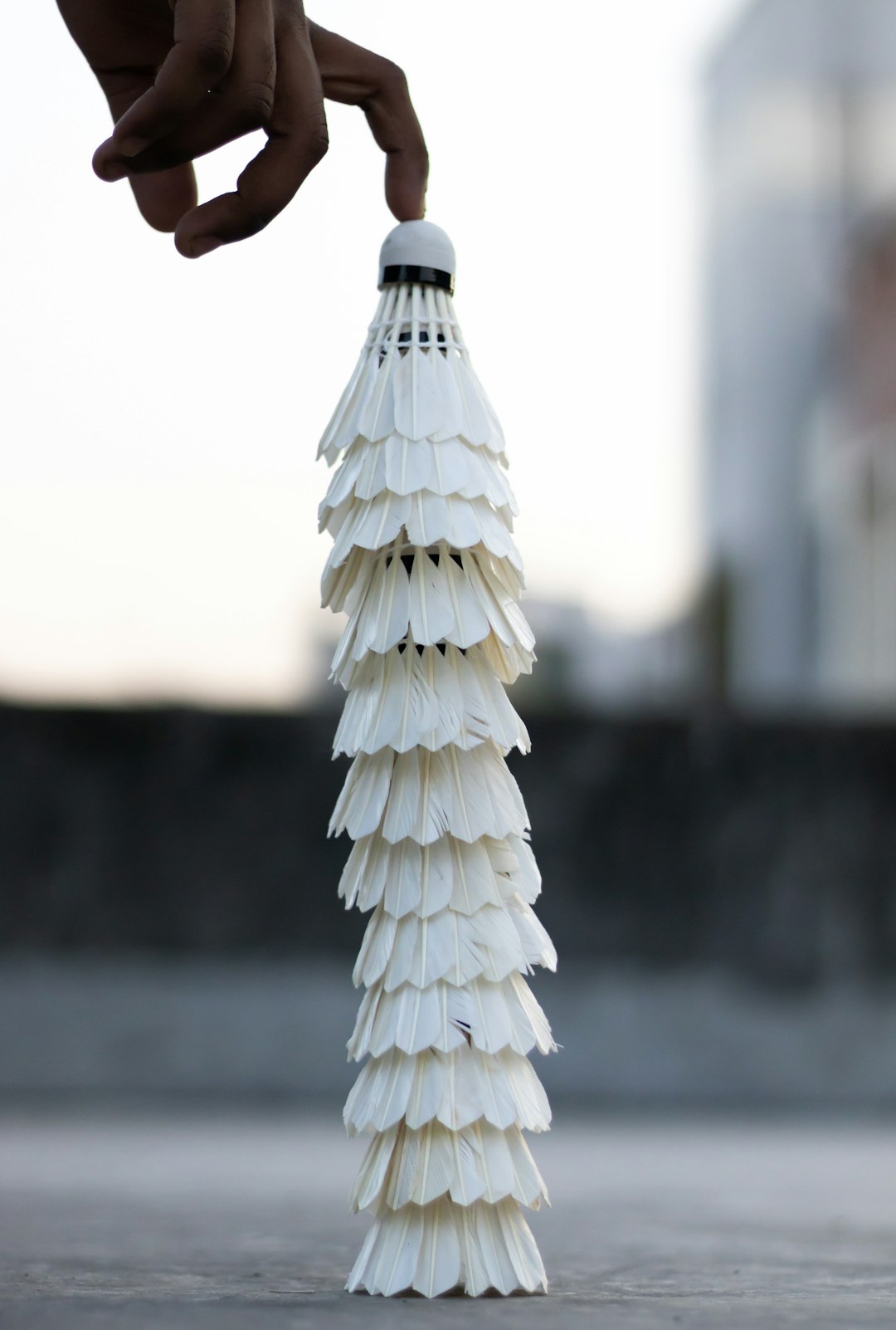 a hand holding badminton shuttlecock, the head of which is made up by many white feathers, stacked on top each other in vertical line to form a mini tower that stands at an angle on its base. The background shows part of a building and sky. The focus is on capturing details such as texture of feather. The overall mood evokes tranquility with minimalism. –ar 43:64