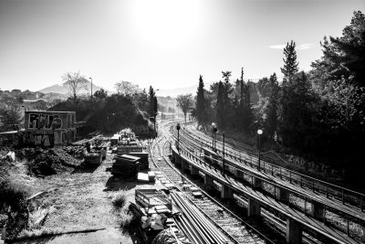 Black and white photography of the construction site for an underground train station in a small town in Greece. On one side there are old railway tracks. Sunlight is shining through trees. In front of us we can see workers working on laying new concrete roads with steel beams. Shot in the style of [Peter Lindbergh](https://goo.gl/search?artist%20Peter%20Lindbergh). Nikon D850 camera with Nikkor 24-70mm f/3 lens. --ar 128:85