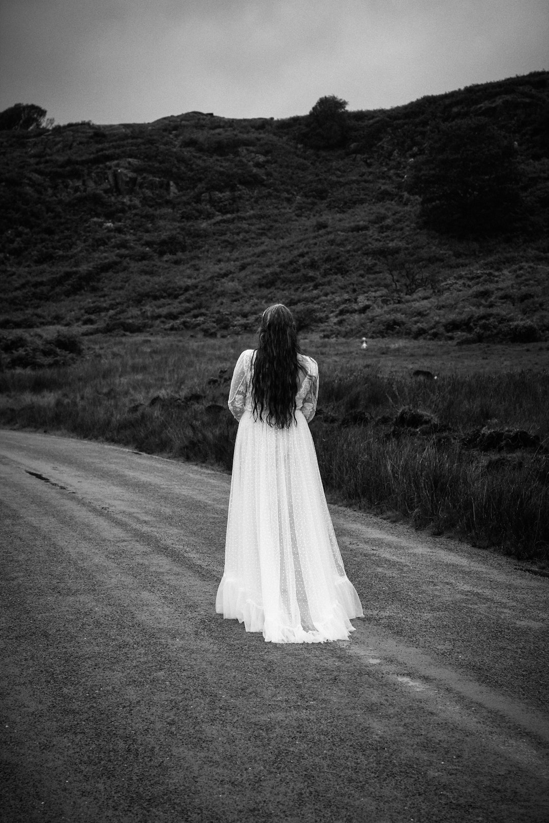 Black and white photography of the back view of an irish woman in her wedding dress standing on side road, plain landscape with hills, lonely vibe –ar 85:128