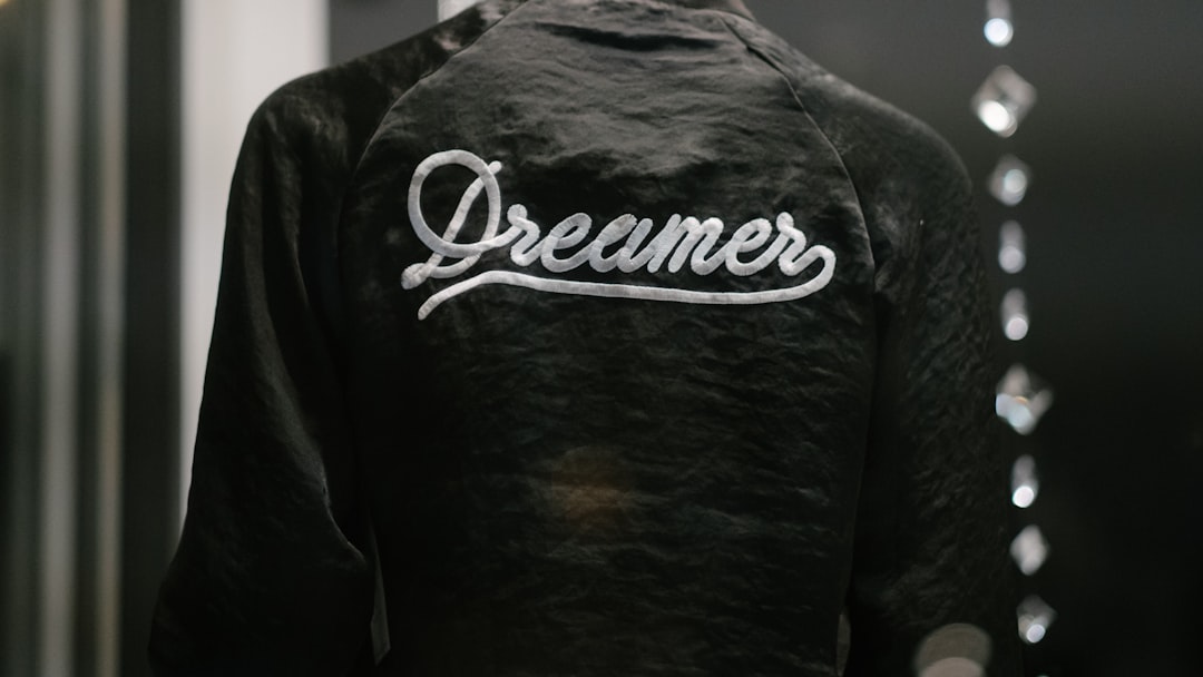 A black baseball jacket with the word “Dreamer” embroidered in white on it, a closeup product shot in front of a mirror, in the style of high fashion, monochromatic shadows, studio photography, soft lighting, hyperrealistic details, luxury brand aesthetics, product design photography, a photo taken from behind the subject. –ar 16:9