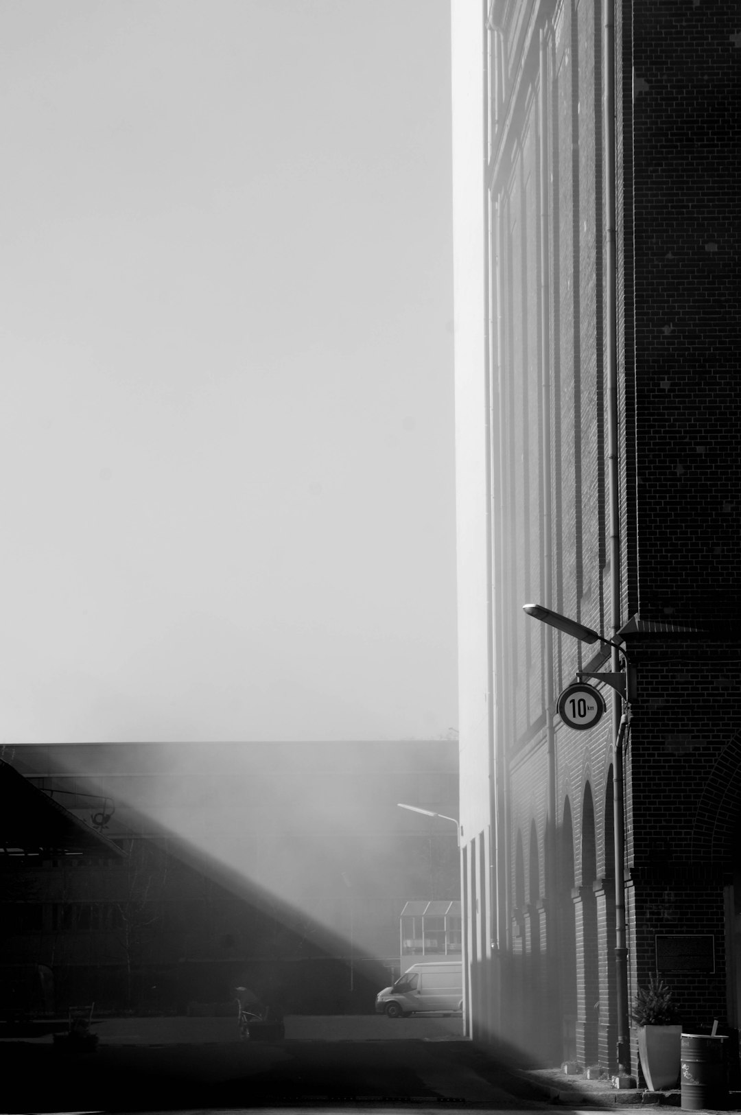 A black and white photograph of the side profile corner of an industrial building with cars parked in front, with sunlight streaming through, and fog on one part of it. There is also a sign pointing to number “40” hanging from above. The style should be minimalist and cinematic, capturing urban architecture in raw photography. –ar 85:128